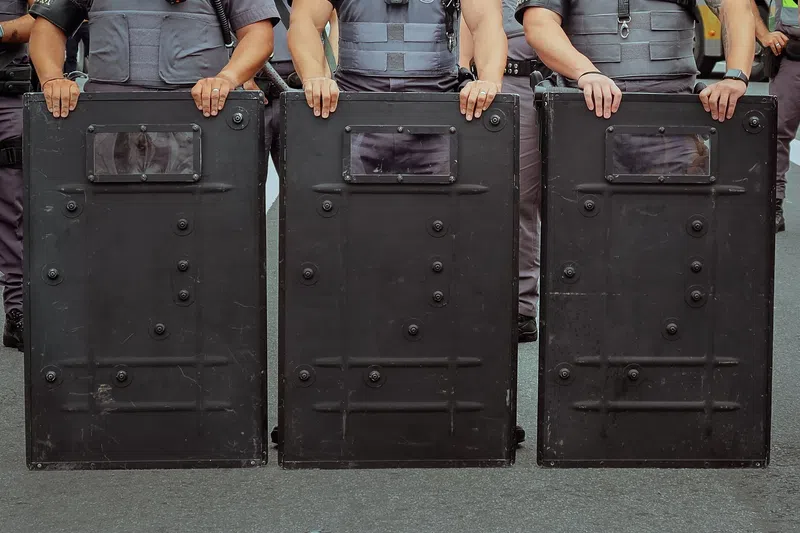 Police officers holding black riot shields in a line during a public safety operation.